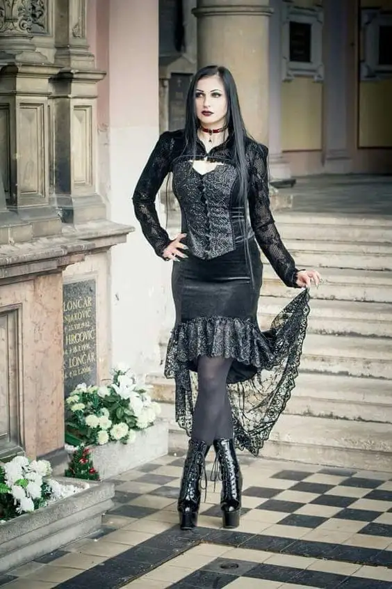 A woman in a black gothic outfit with lace, corset, and high boots poses confidently in front of a cemetery with tombstones…