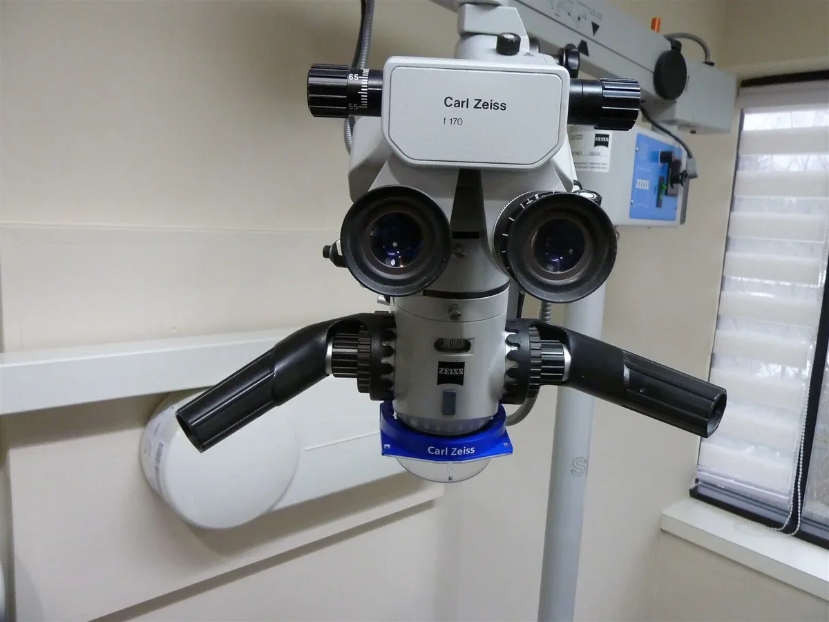 A close-up photo of a Carl Zeiss surgical microscope with two eyepieces and black adjustment knobs, mounted on a stand in a…