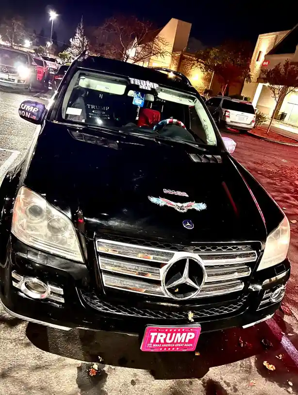 A black Mercedes-Benz SUV is parked at night in a lot, covered in Trump 2020 and Make America Great Again political stickers…