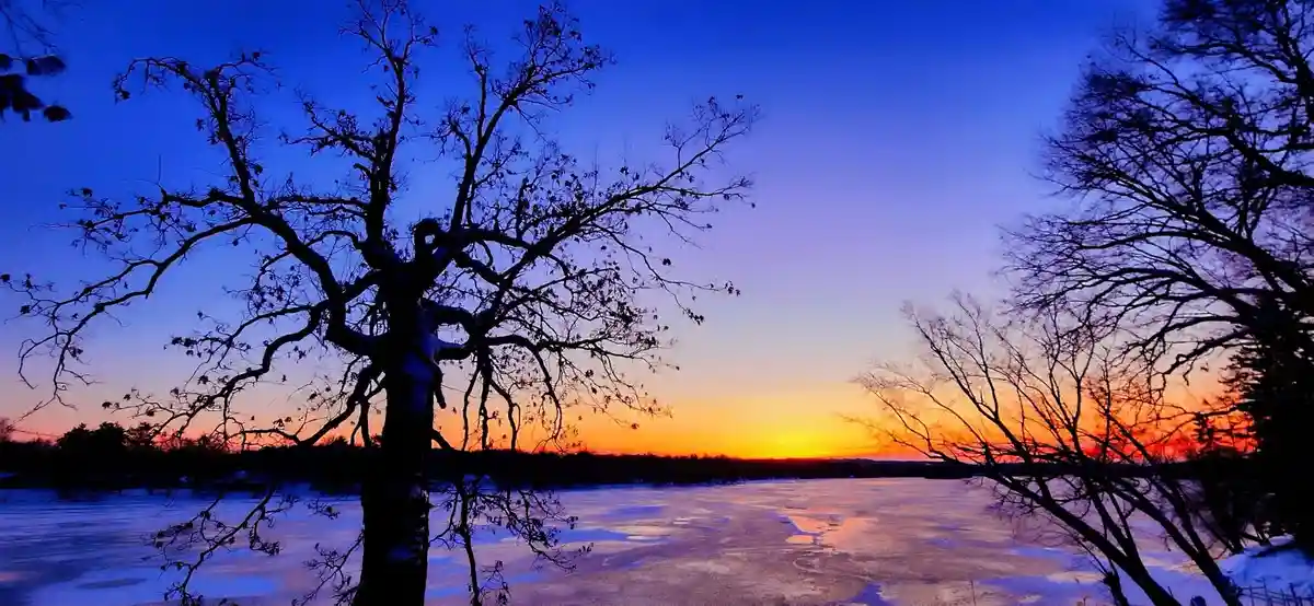A serene winter sunset over a frozen lake, with bare trees silhouetted against a gradient sky of deep blue, purple, and…