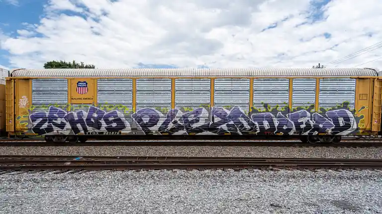 A yellow Union Pacific freight train car with a large purple and yellow graffiti tag reading 'this one's dead' across its…