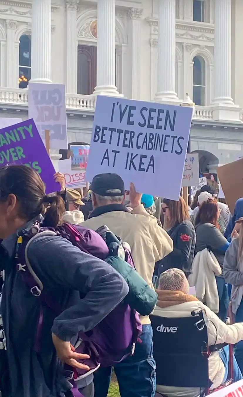 A crowd of protesters holds signs outside a grand white building with columns. One man in a beige jacket and black cap…
