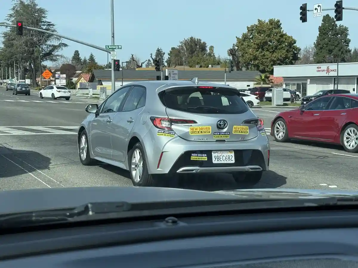 A silver Toyota Corolla is stopped at a red traffic light with multiple yellow 'NEW DRIVER PLEASE BE PATIENT' stickers on…
