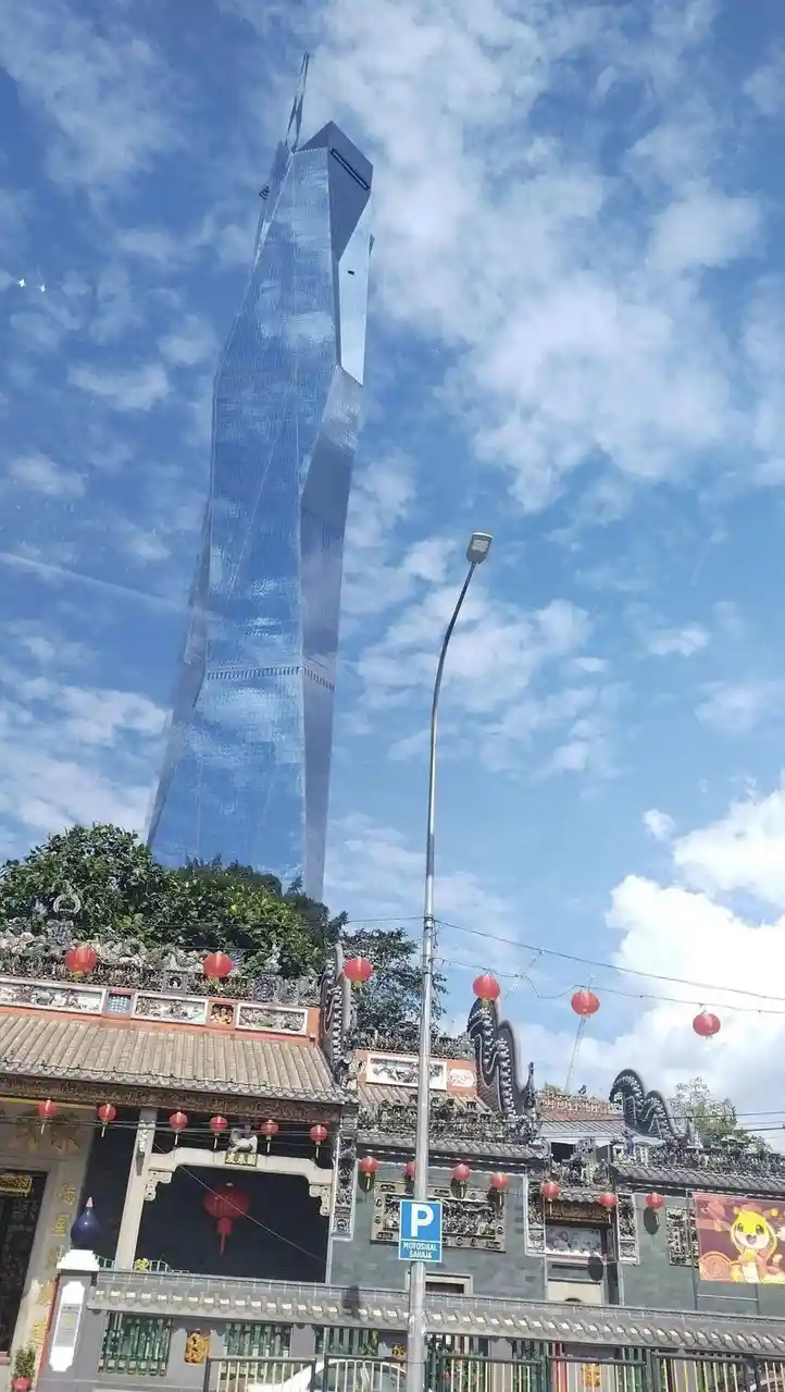 A tall, modern glass skyscraper with a distinctive twisted design towers over a traditional ornate temple adorned with red…