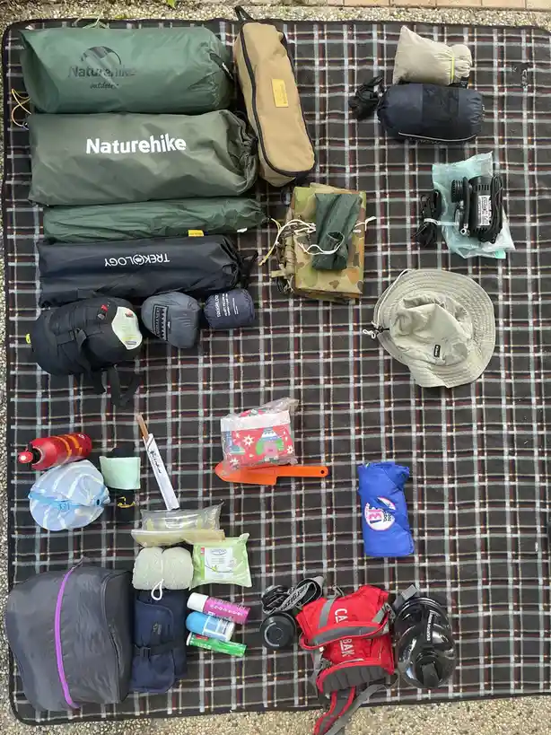 An overhead shot displays a meticulously organized collection of camping gear and personal items spread across a plaid…