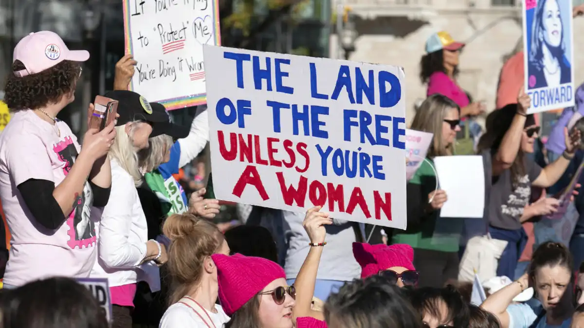 A crowd of protesters at an outdoor rally holds handmade signs; the central sign reads 'THE LAND OF THE FREE UNLESS YOU'RE A…