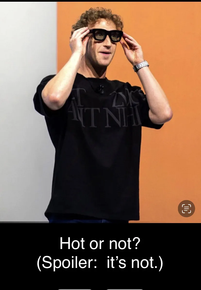 A man with curly hair wearing a black t-shirt and large black glasses is adjusting them with both hands while standing in…