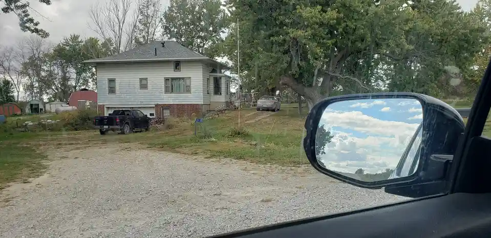 A photo taken from inside a car shows a rural house with a gravel driveway, a black pickup truck parked nearby, and a large…