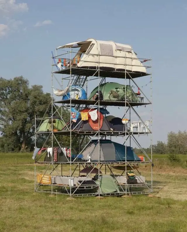 A tall, multi-level metal scaffolding structure stands in a grassy field under a blue sky, with various tents and camping…
