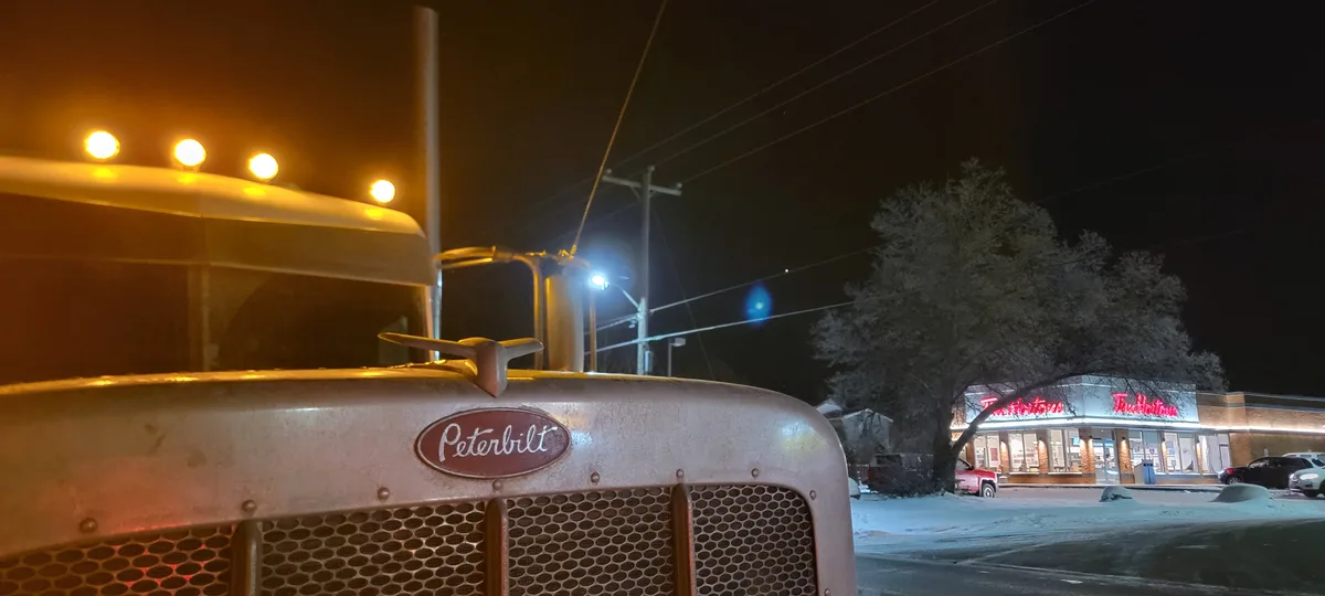 A close-up of the front of a vintage Peterbilt truck at night, its headlights glowing, parked in front of a Tim Hortons with…