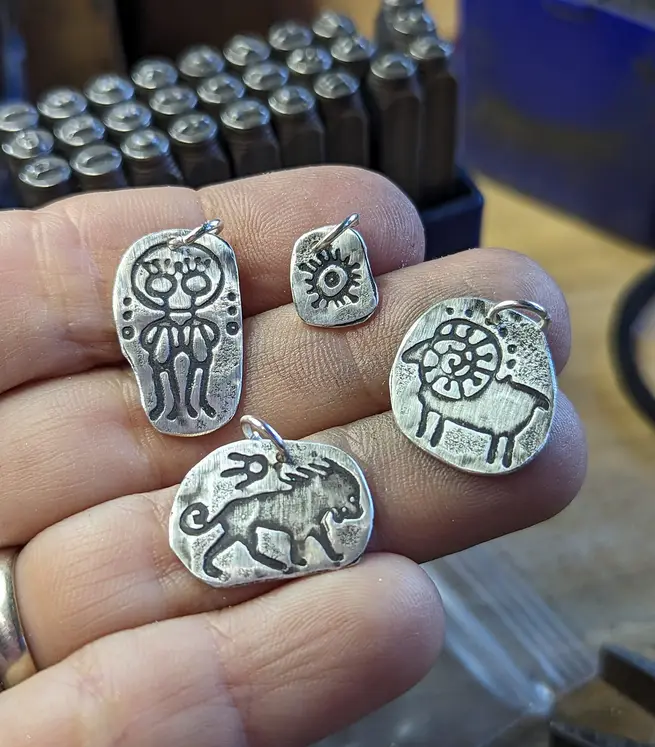 A hand holds four small, silver-colored metal pendants with stylized ancient-looking designs: a humanoid figure, a sun-like…