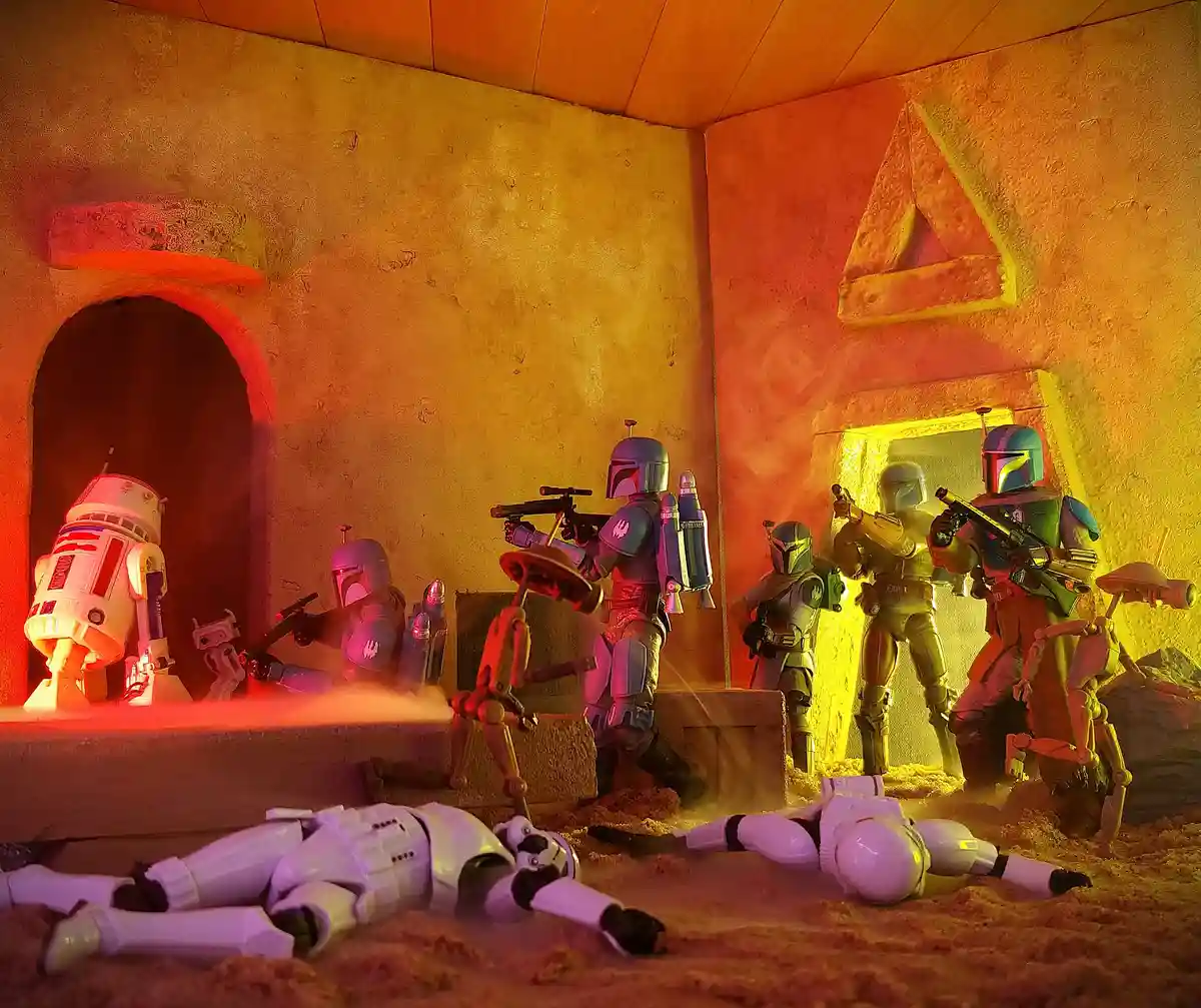 A diorama scene featuring Star Wars action figures: a droid and Mandalorian warriors in a desert-like room with two fallen…