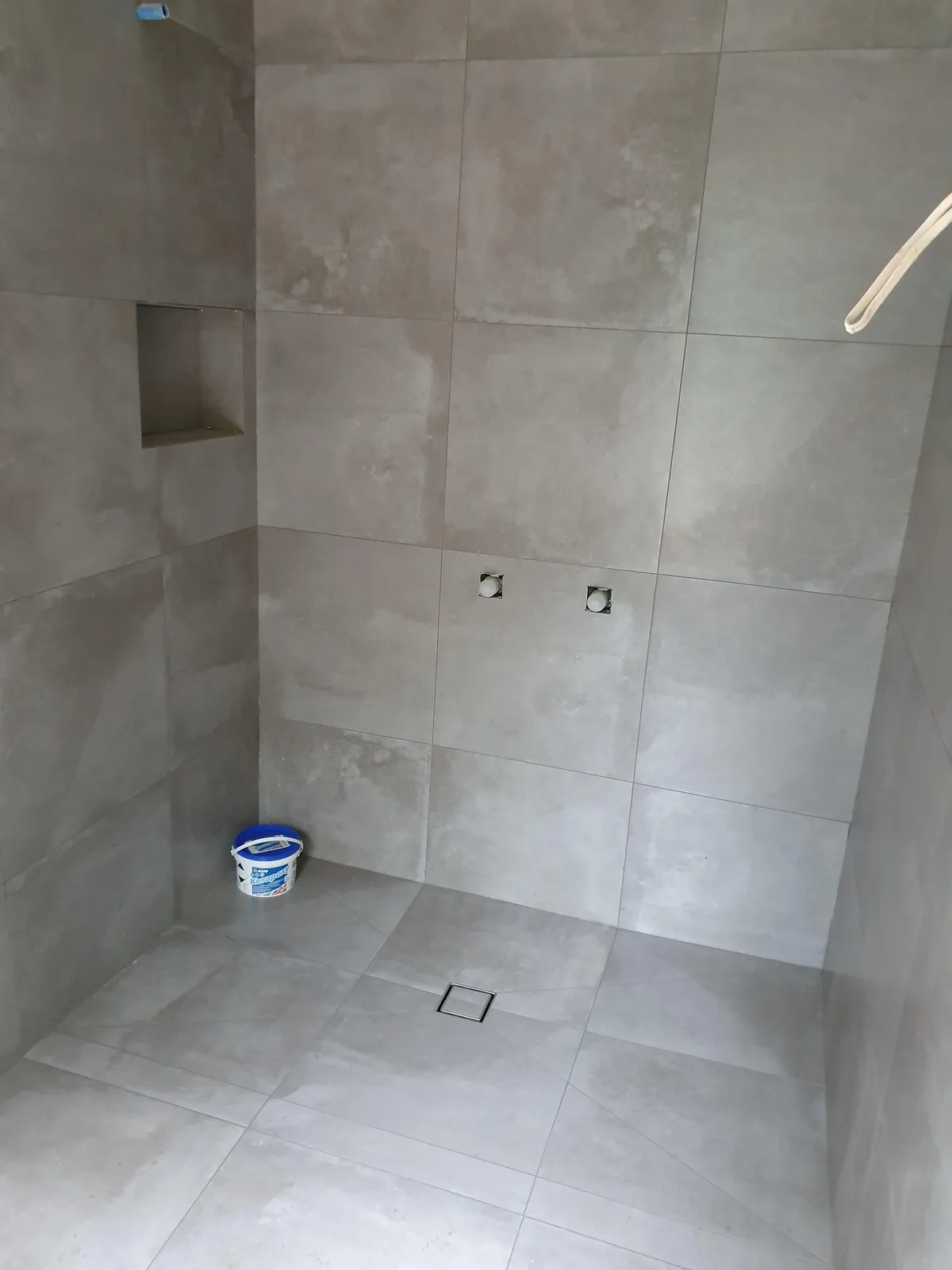 An empty, tiled shower stall with gray tiles, a floor drain, and two exposed plumbing holes on the wall. A small recessed…