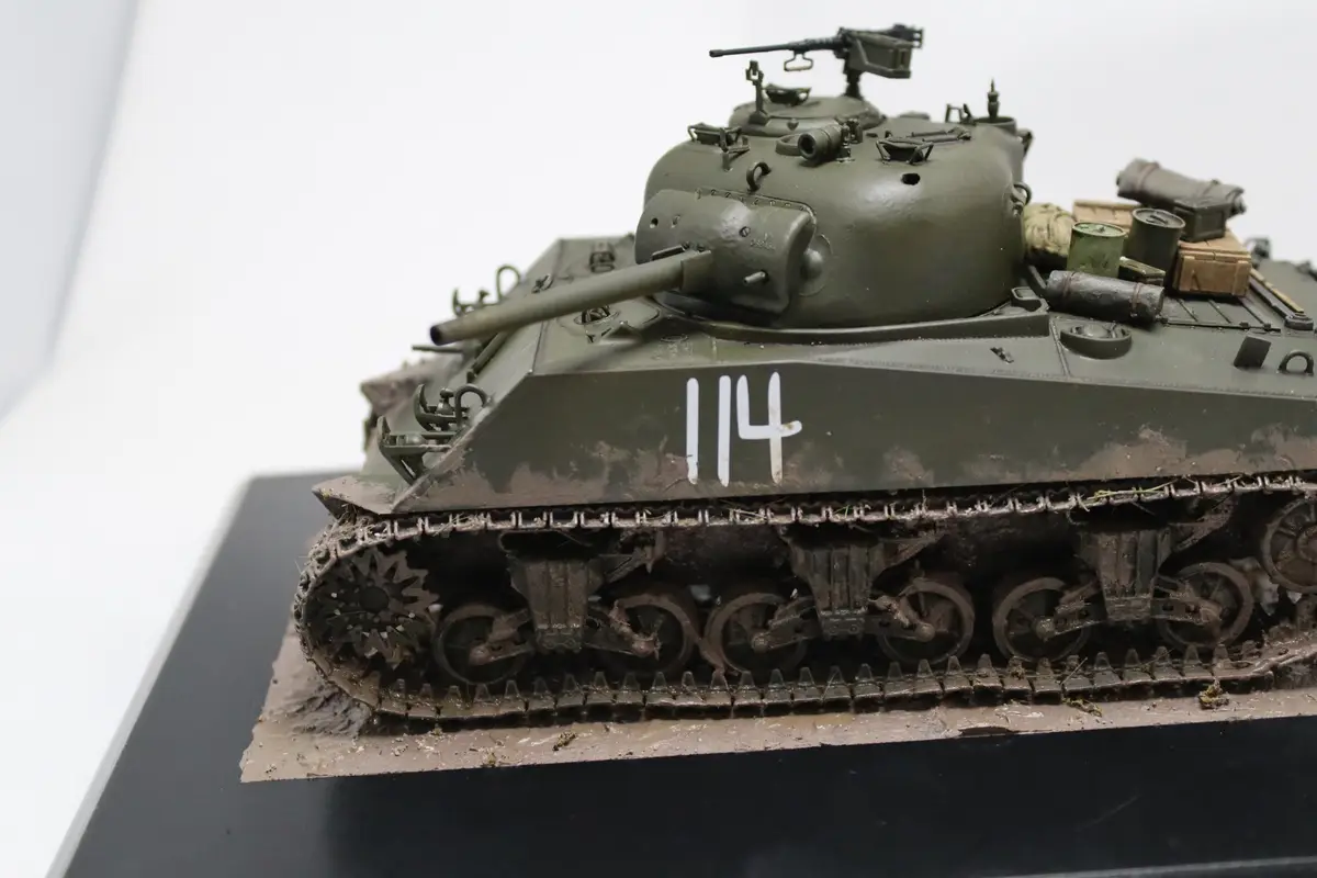 A detailed scale model of a green military tank, marked with the number 114 on its side, sits on a muddy diorama base. The…