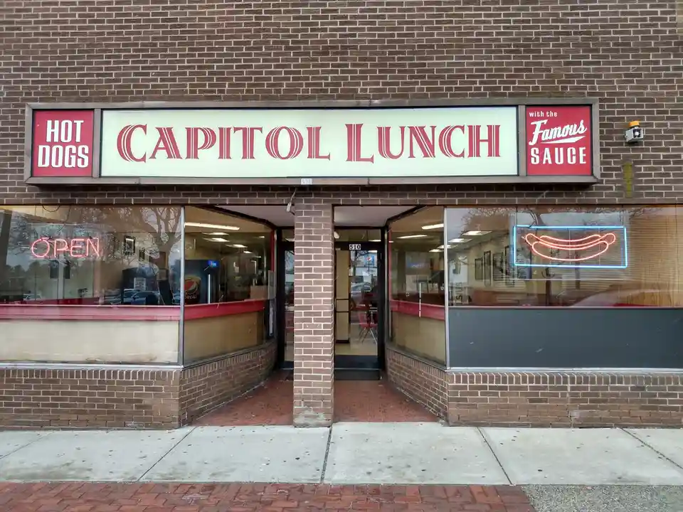 A storefront of a brick building with a large sign reading 'CAPITOL LUNCH' flanked by 'HOT DOGS' and 'with the Famous SAUCE'…