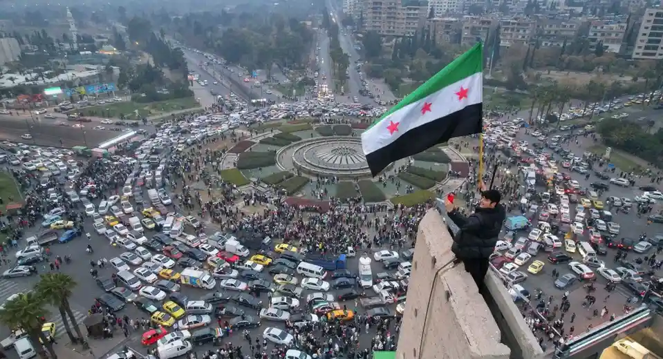A man stands atop a concrete structure overlooking a massive protest scene filled with crowds and traffic, holding a Syrian…