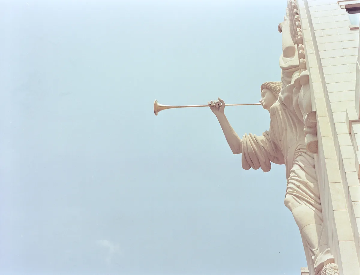 A stone statue of a person holding a trumpet to their lips, mounted on the corner of a building against a clear blue sky…