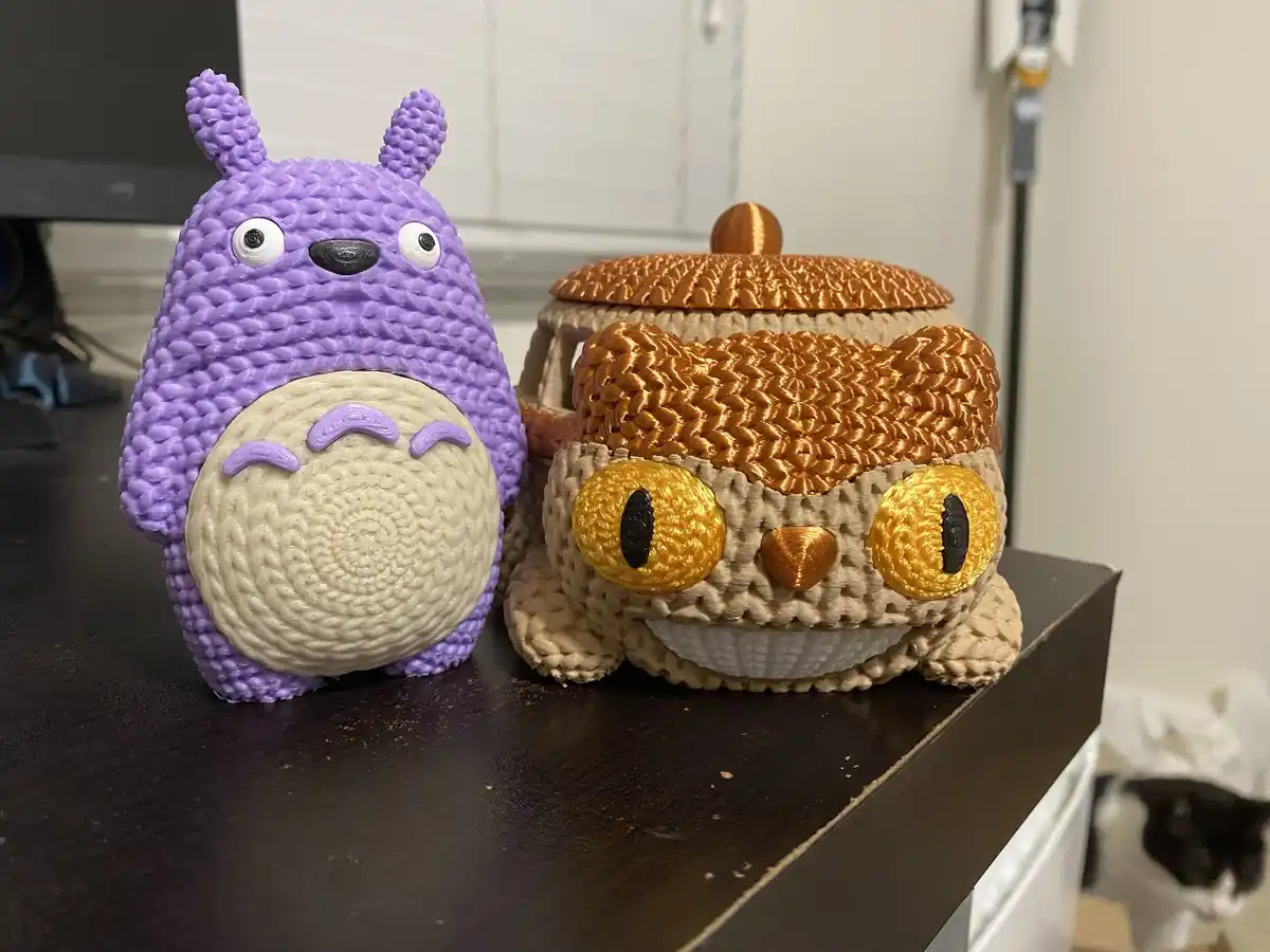Two 3D-printed, crocheted-style figurines sit on a dark wooden surface: a purple Totoro with a cream belly and a tan…