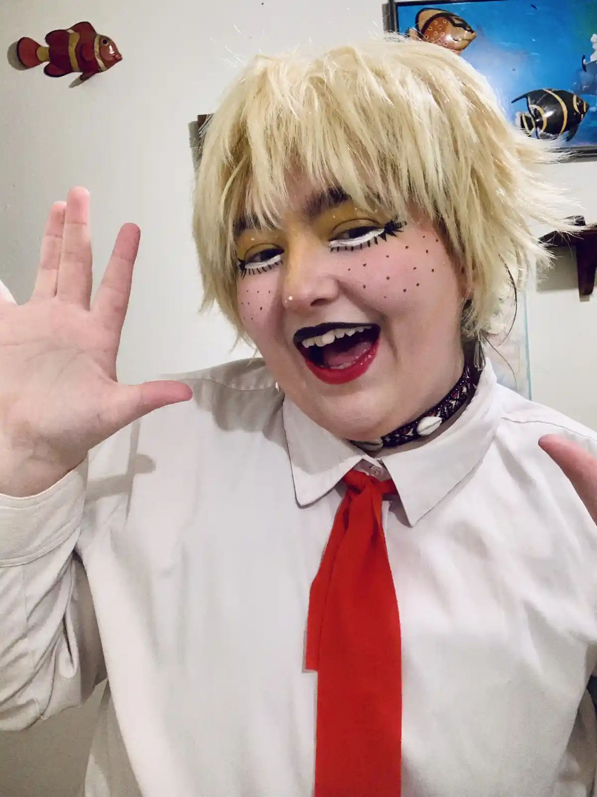 A person with a blonde wig and exaggerated makeup, including black eyelashes, yellow eyeshadow, and freckle-like dots on…