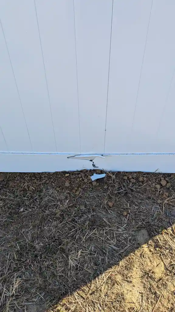 A close-up photo shows a white vinyl siding wall with a jagged, torn section at the bottom where the siding meets the…