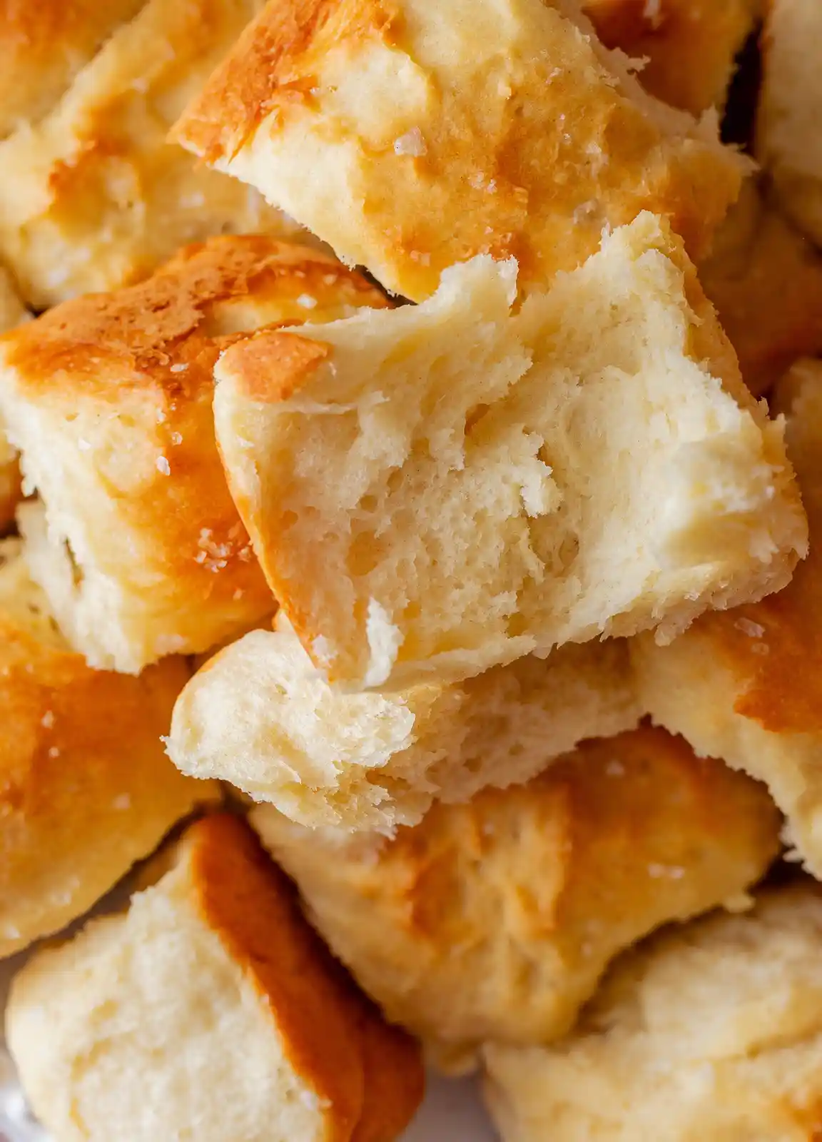 A close-up photo shows golden-brown, soft-looking bread rolls piled together, with one roll broken open to reveal its airy…