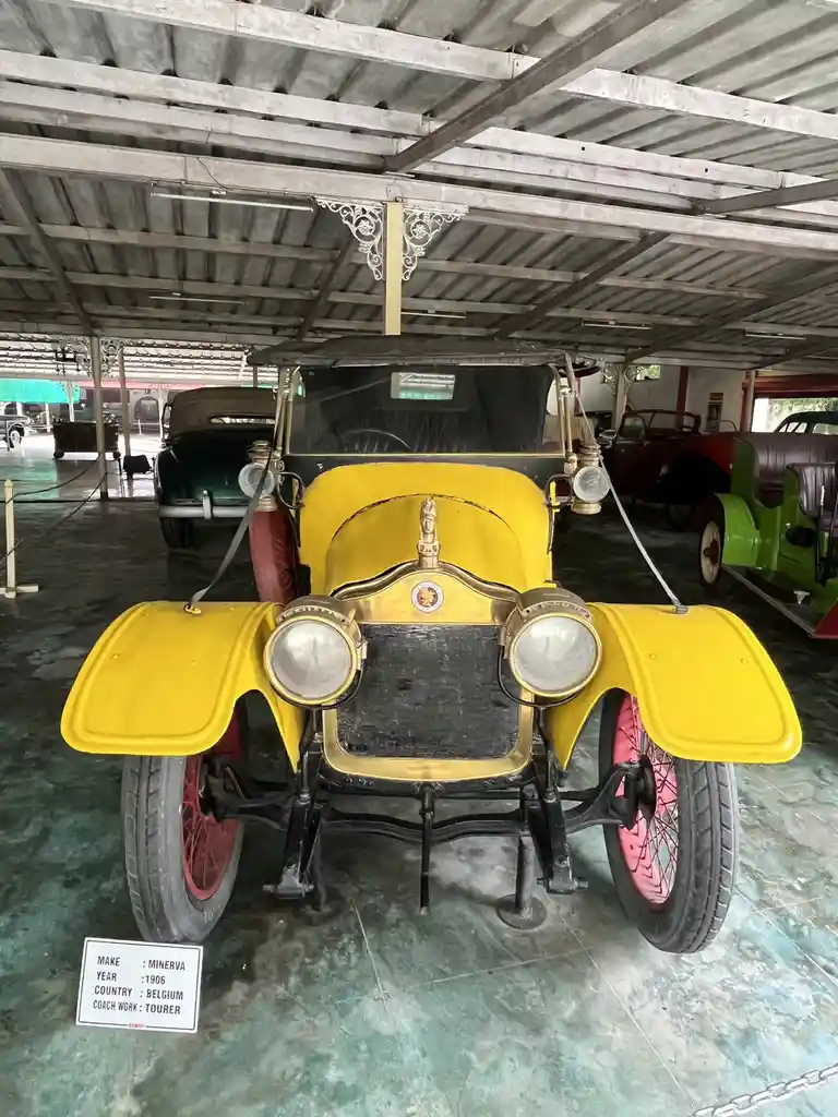 A bright yellow 1906 Minerva Tourer from Belgium is displayed in a covered car museum, featuring large round headlights, red…
