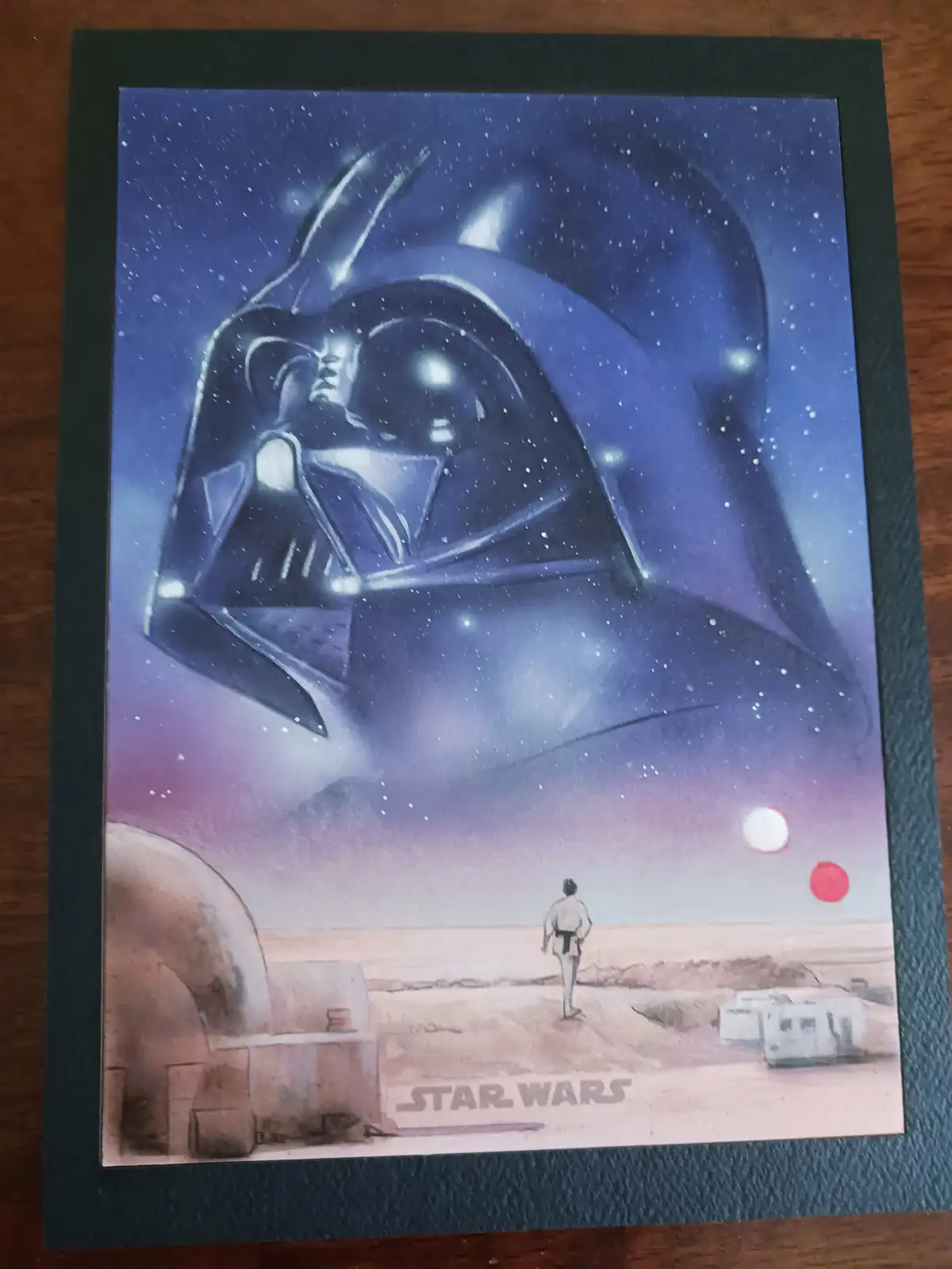 A Star Wars-themed artwork depicts a lone figure in a desert landscape, looking up at a massive, looming Darth Vader helmet…