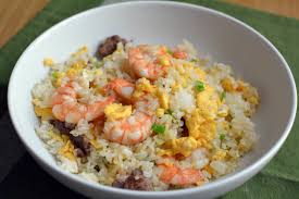 a close-up photo of a white bowl filled with fried rice, visible shrimp, scrambled egg, and small green peas, resting on a…