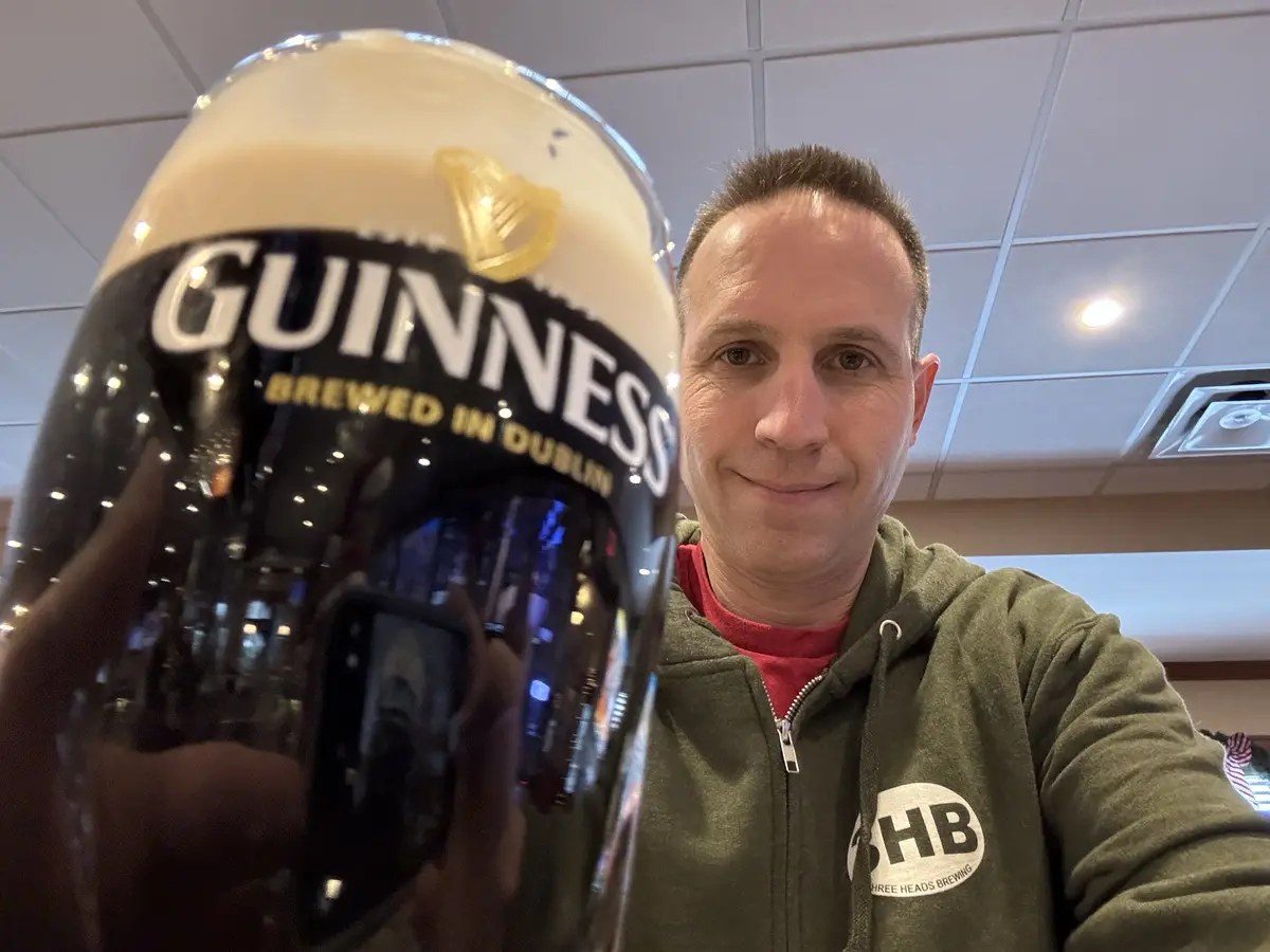 A man in a green hoodie takes a selfie holding a large, frothy glass of Guinness beer, which dominates the foreground. The…
