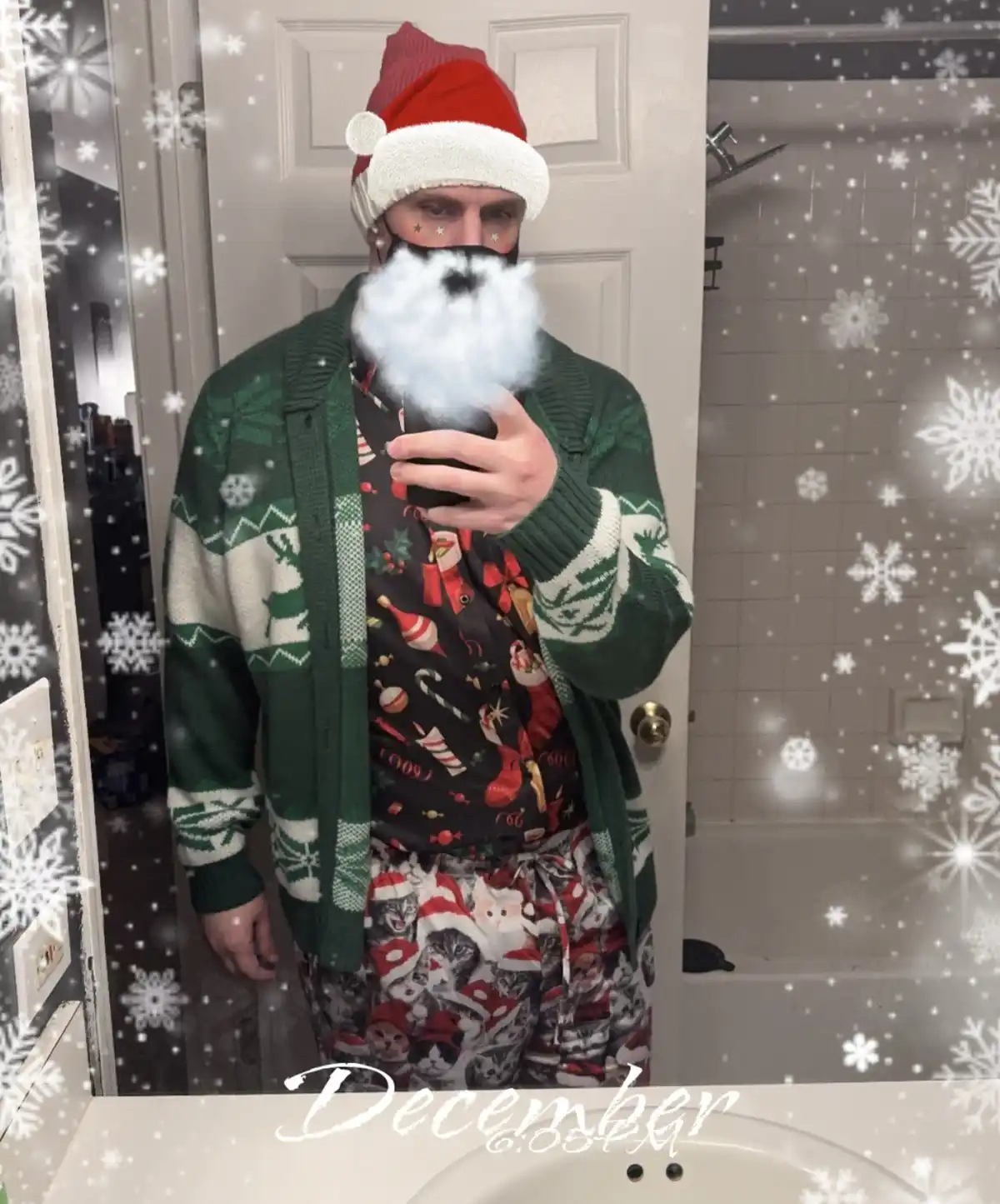 A person in a bathroom mirror selfie wears a red Santa hat, a green holiday sweater, and pajamas with cats in Santa hats…