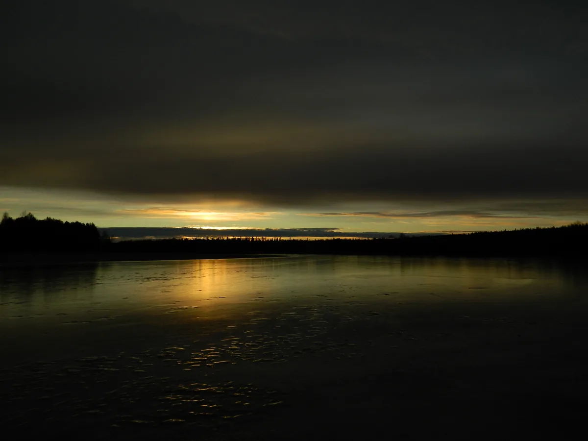 A serene, dimly lit landscape photo shows a frozen lake reflecting a low, golden sunset through dark clouds, with…