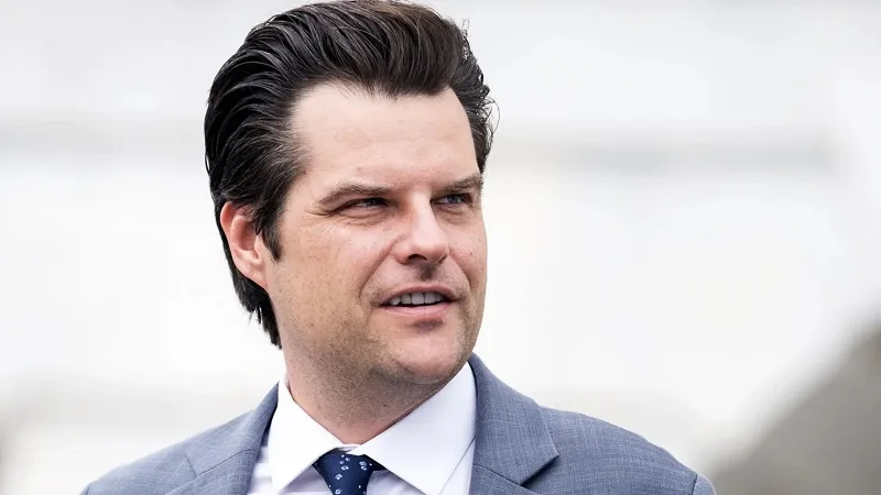 a man with dark hair styled back, wearing a gray suit, white shirt, and patterned tie, looks slightly to his right with a…