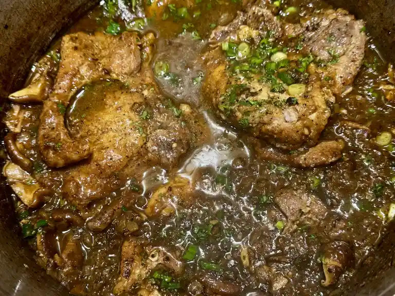 A close-up photo of a simmering stew in a dark pot, featuring chunks of cooked chicken, sliced mushrooms, and a rich brown…