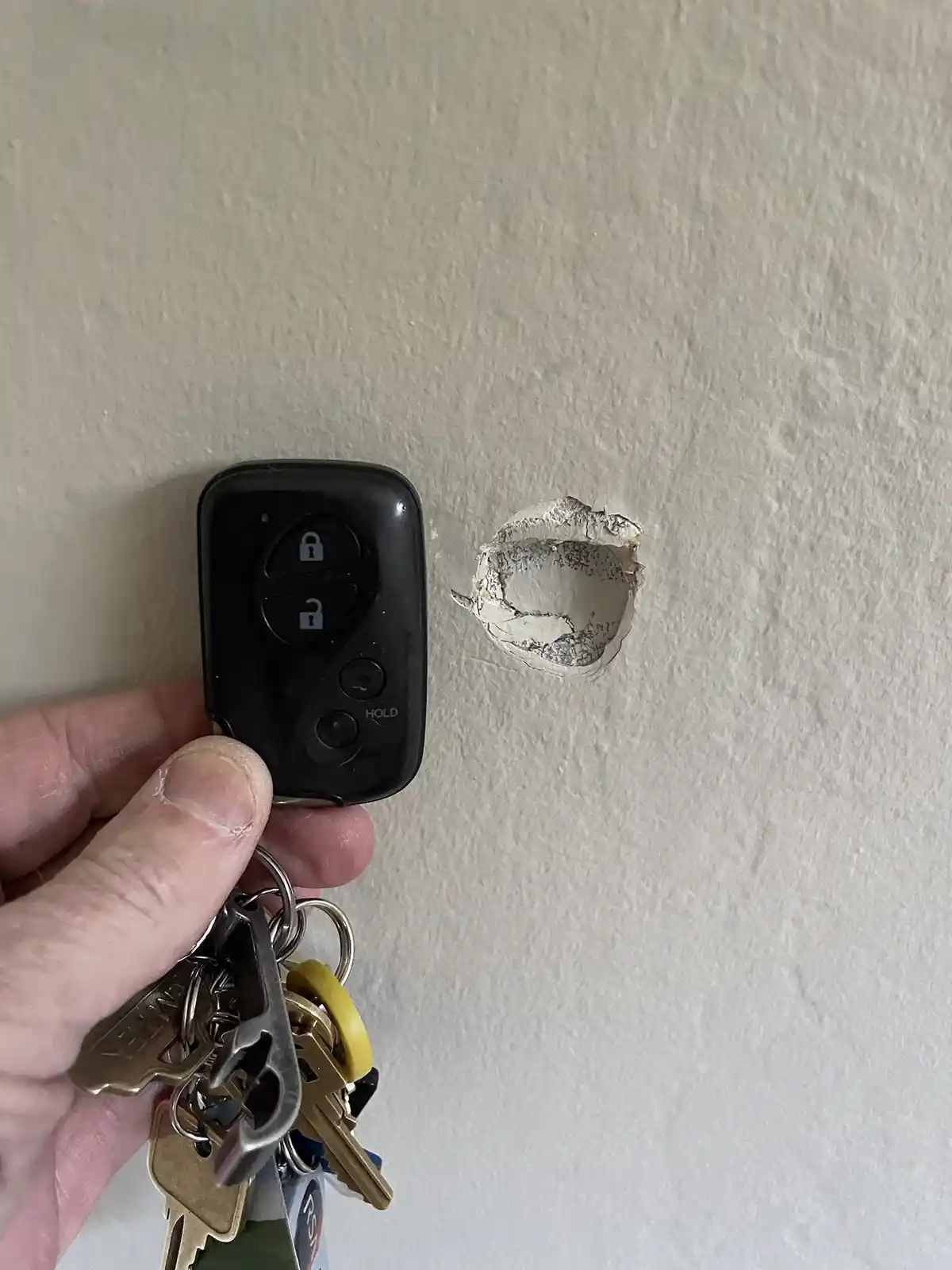 A hand holds a black car key fob with lock/unlock buttons and a keychain with multiple keys next to a circular hole punched…
