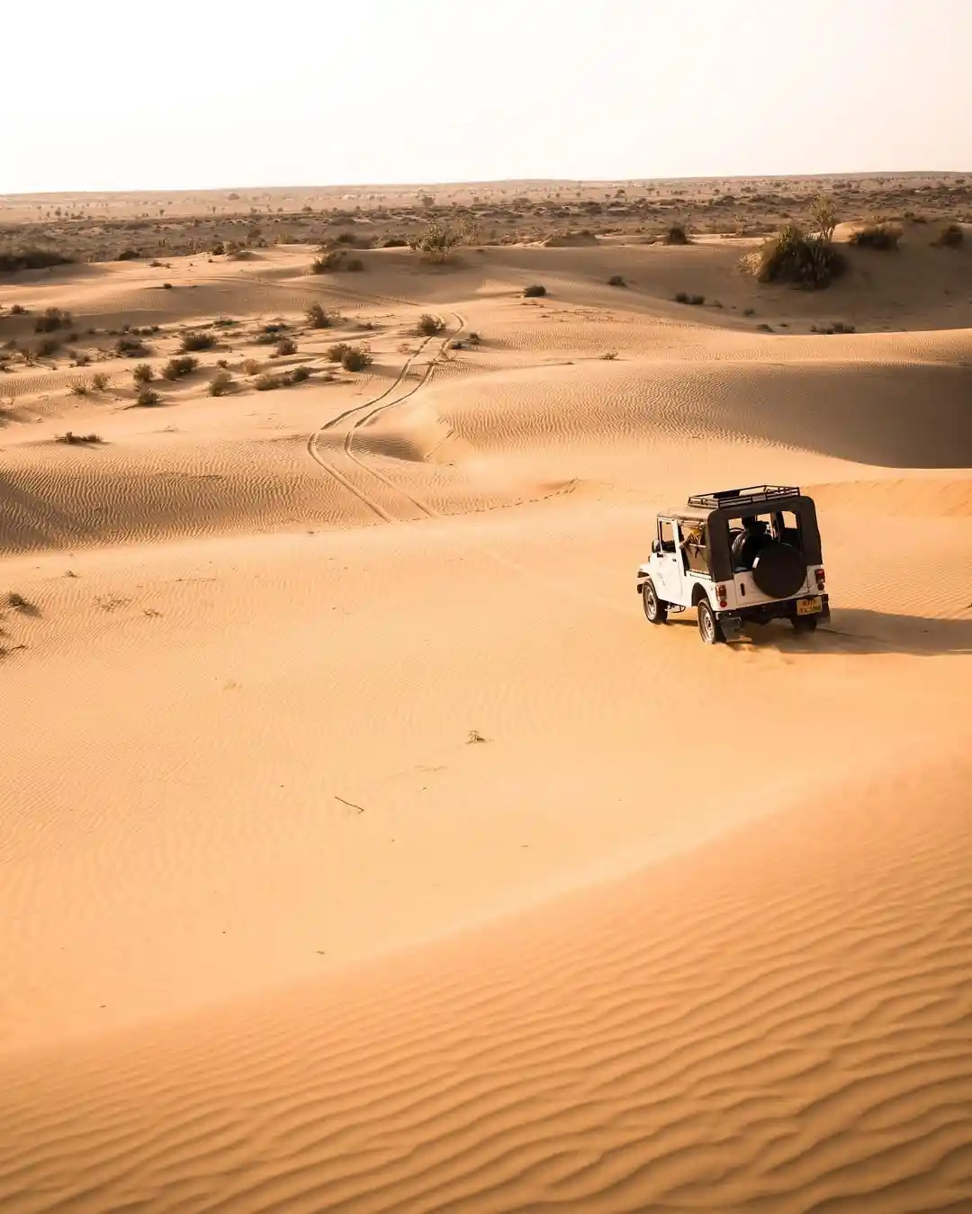 A white off-road vehicle drives across a vast, rippled desert landscape with dunes stretching to the horizon. The vehicle…