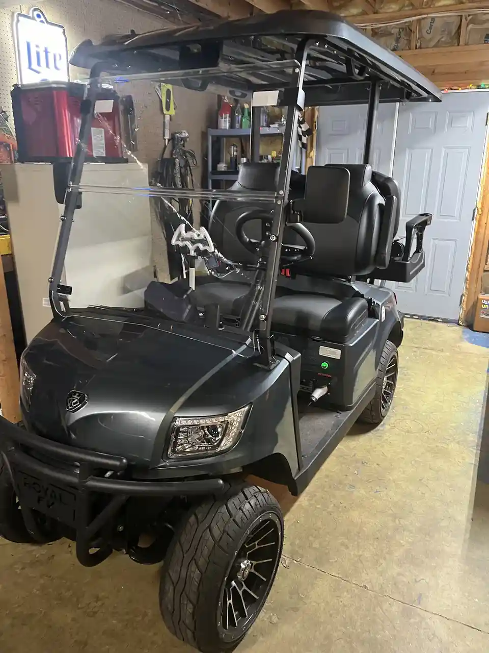 A customized black golf cart with a Batman logo steering wheel is parked in a garage. The cart has large off-road tires, a…