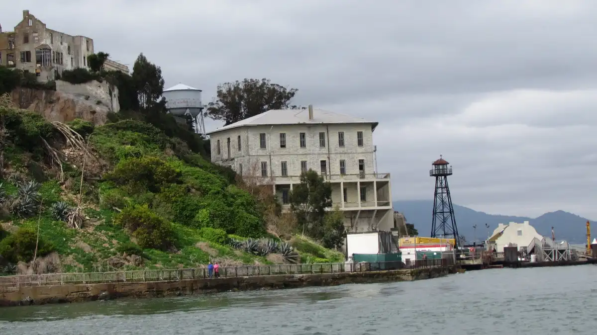 A wide shot of Alcatraz Island under an overcast sky, showing its historic stone buildings, a water tower with faded text…