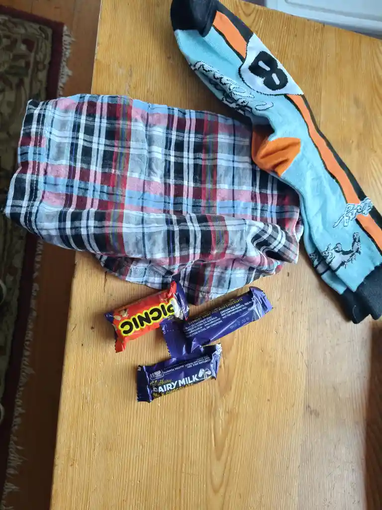 A folded plaid shirt and a colorful cartoon-patterned sock lie on a wooden table next to three chocolate bars: one…
