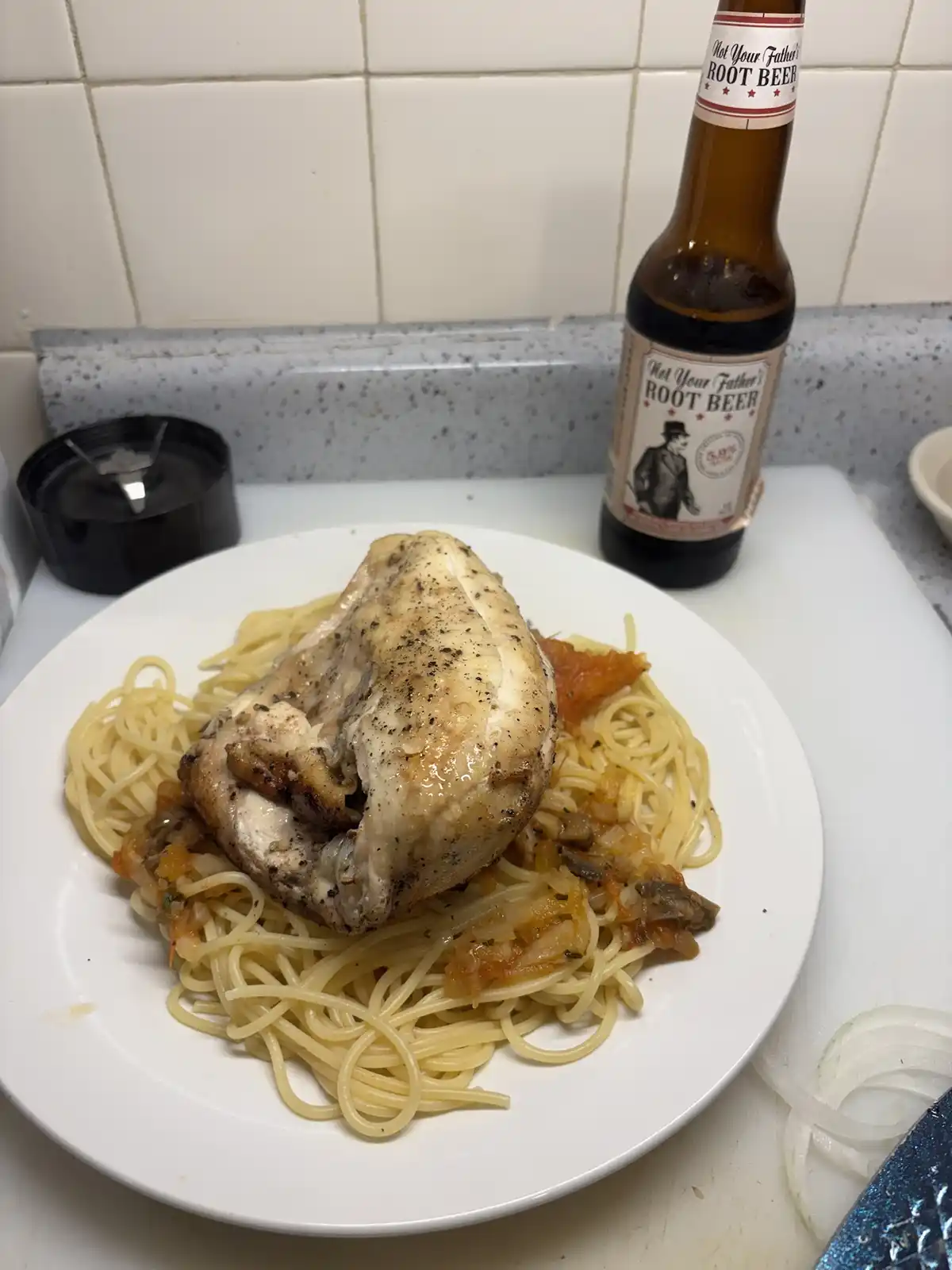 A plate of spaghetti topped with a large, seasoned chicken breast sits on a kitchen counter next to a bottle labeled 'Not…