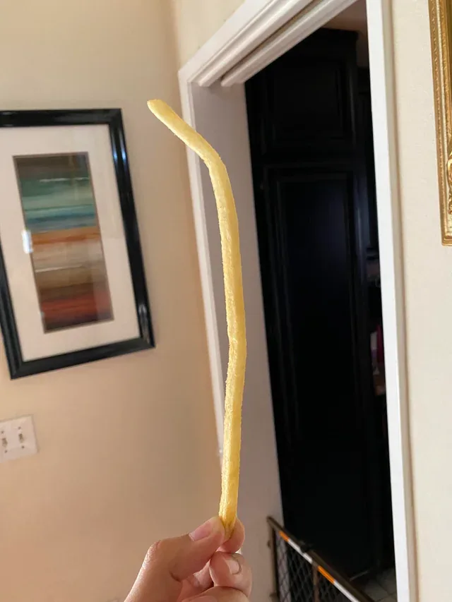 a hand holds up an extremely long, curved french fry against an indoor background with a doorway and framed art on the wall…