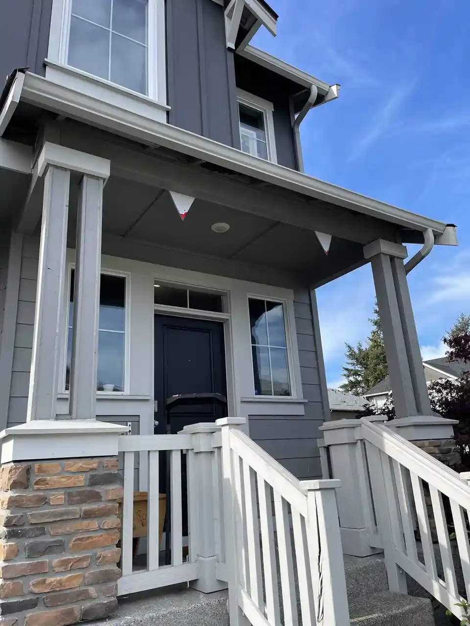 A photo of a modern two-story house with gray siding, white trim, and a white porch railing under a clear blue sky. Two…