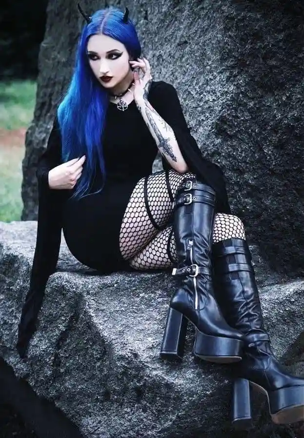 A person with long blue hair and small black horns sits on a large rock wearing a black dress, fishnet stockings, and tall…