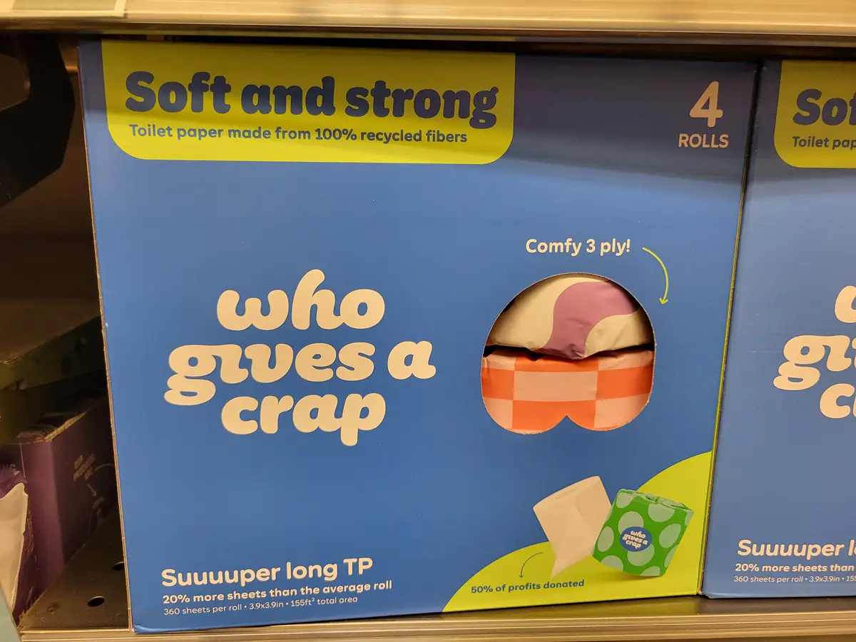 A close-up of a blue 'who gives a crap' toilet paper box on a store shelf, featuring text like 'Soft and strong', 'Comfy 3…