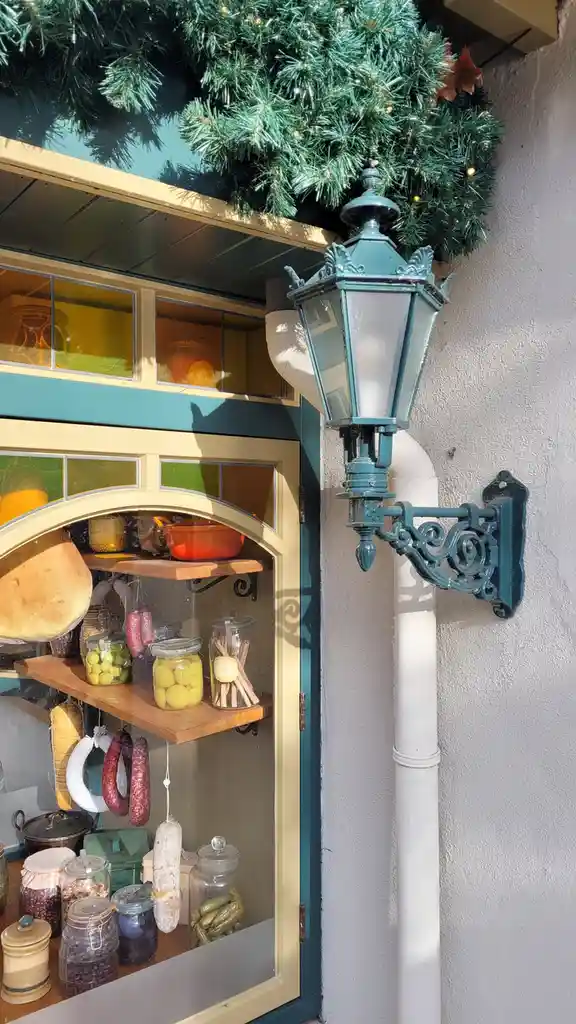 A festive outdoor display features a vintage teal street lamp mounted on a white pipe beside a shop window filled with jars…