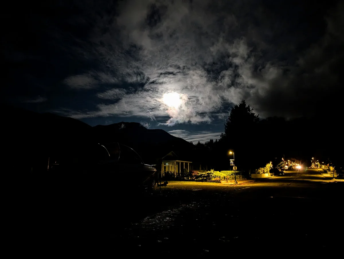 A nighttime photo shows a bright moon partially obscured by wispy clouds above a dark landscape with mountains, a house, and…