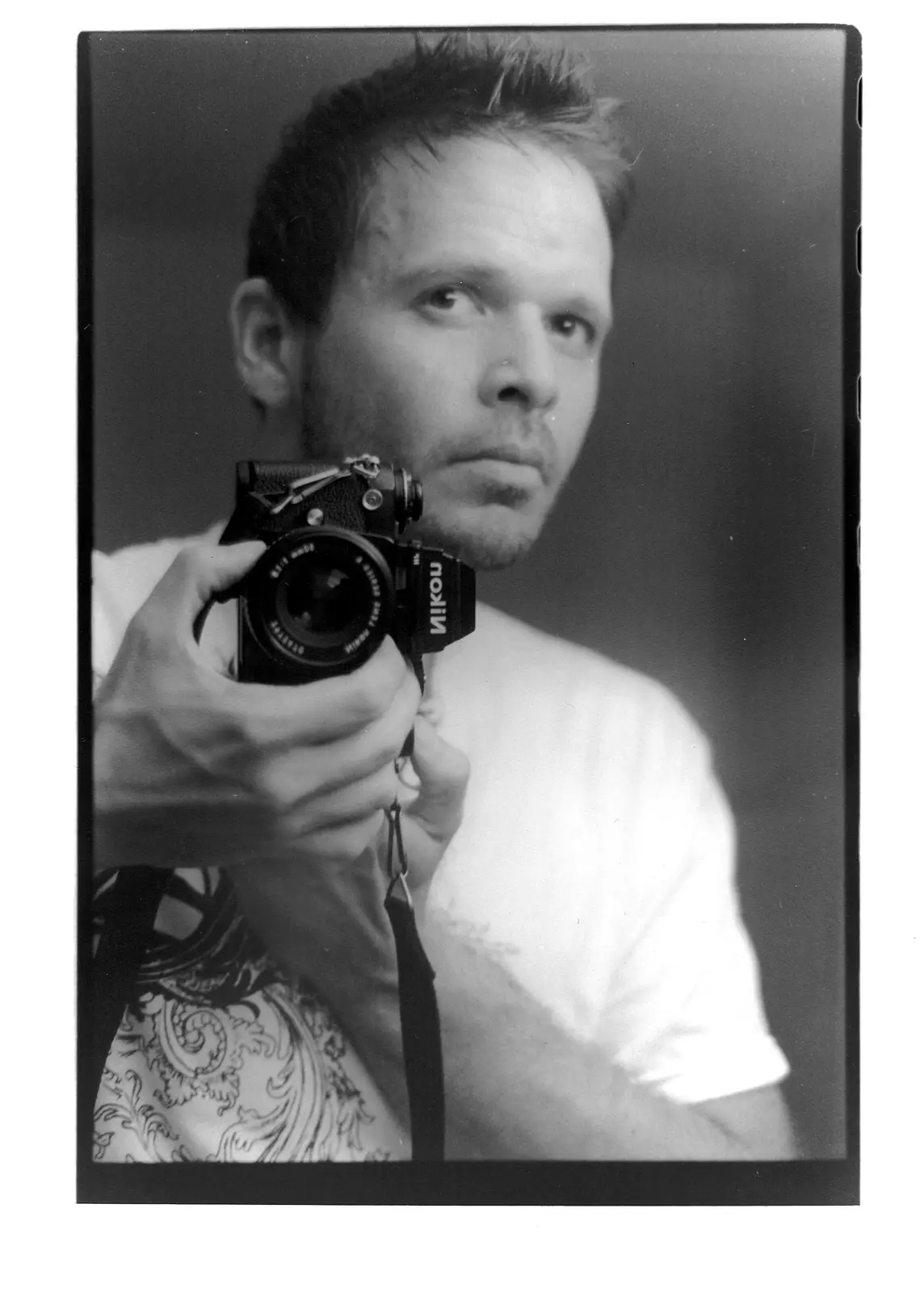 A black-and-white photo shows a man holding a Nikon camera, looking off to the side with a serious expression. He wears a…