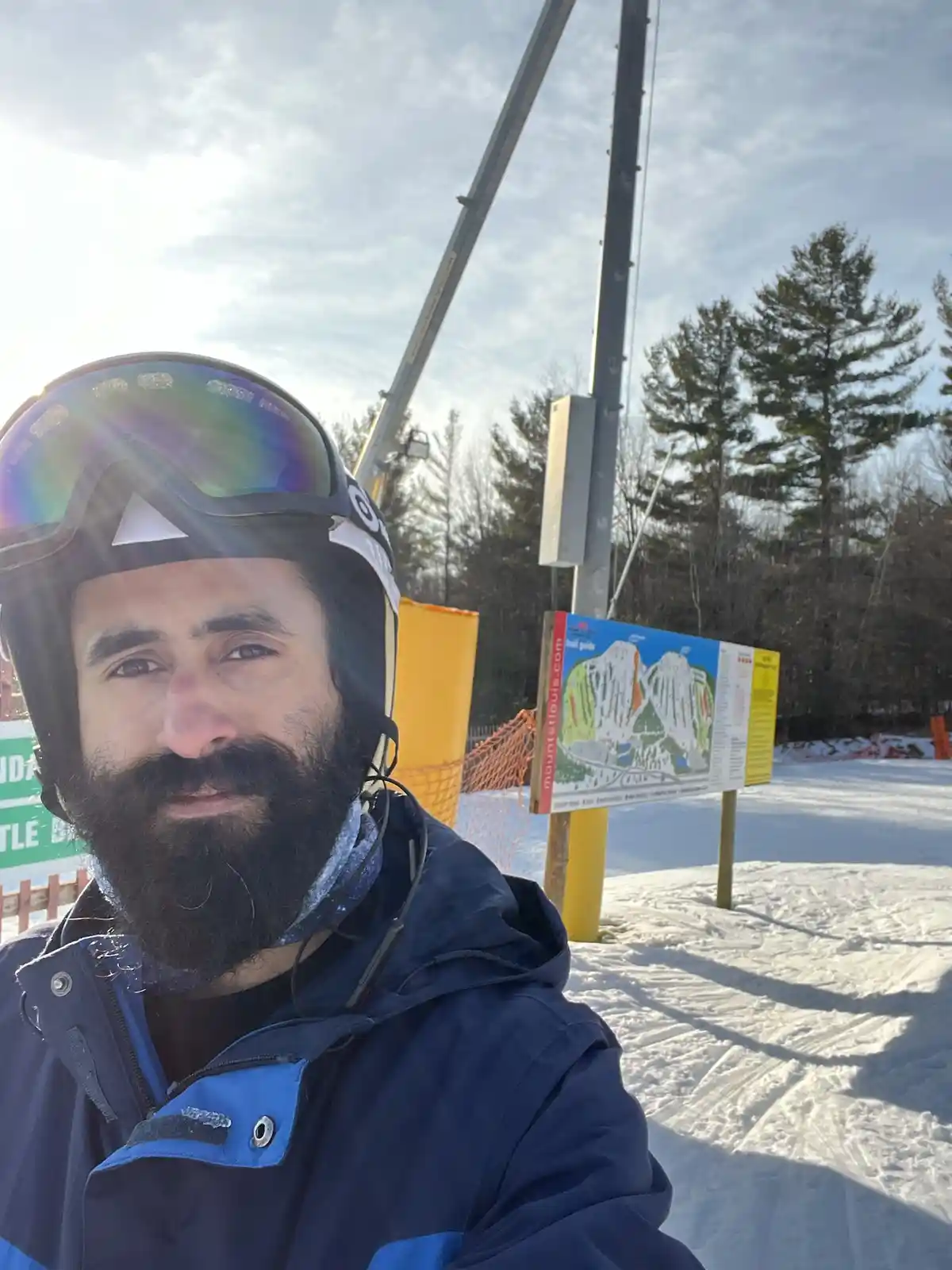 A man with a beard wearing a ski helmet and goggles takes a selfie at a snowy ski resort. Behind him is a trail map sign for…