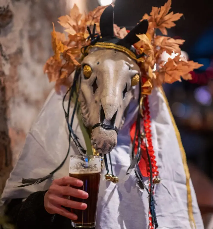 A person wearing a horse skull mask adorned with autumn leaves and decorative cords holds a pint of dark beer. The mask has…