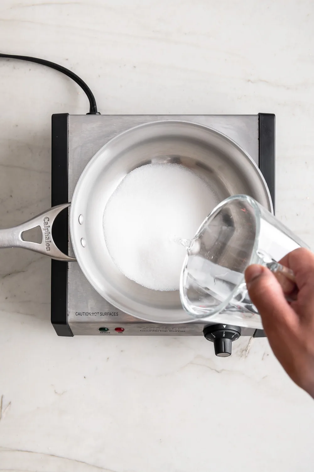 A hand pours water from a glass measuring cup into a stainless steel saucepan containing granulated sugar, which sits on a…