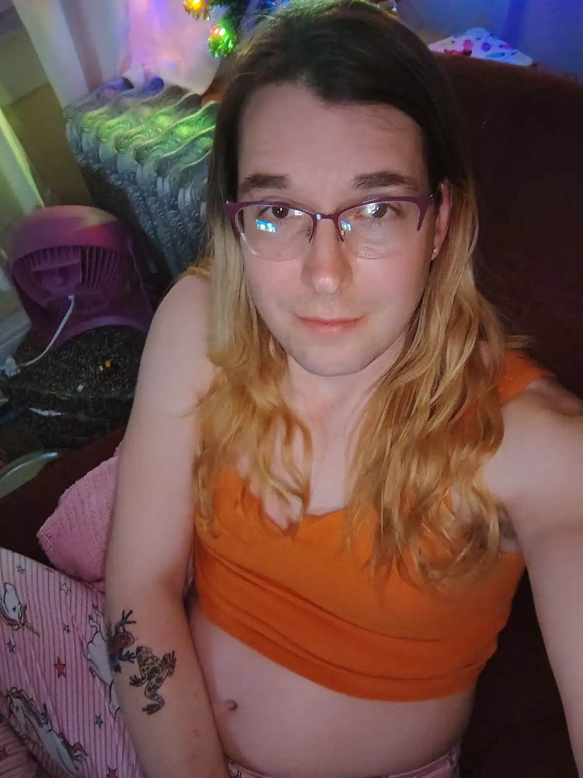 A person with long blonde hair and purple glasses sits on a couch wearing an orange crop top, looking at the camera with a…